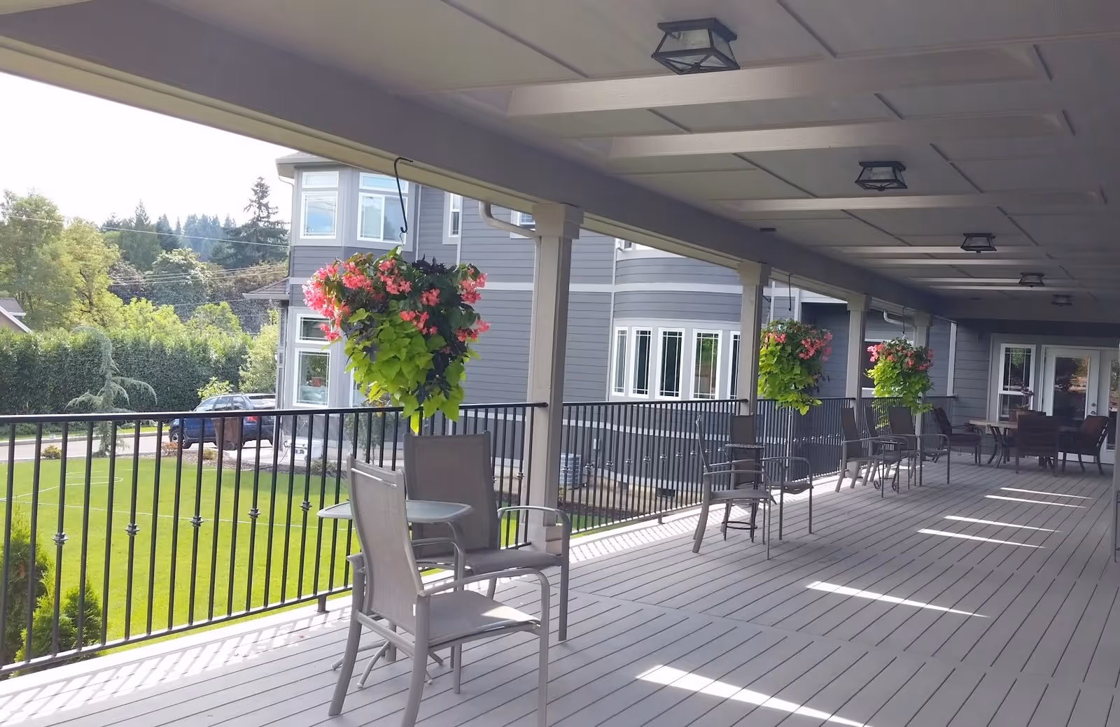 Covered outdoor patio area with several chairs and small tables arranged along a railing. Hanging flower baskets with pink and green foliage are suspended from the ceiling. The patio overlooks a green lawn and trees, with a gray building visible in the background.