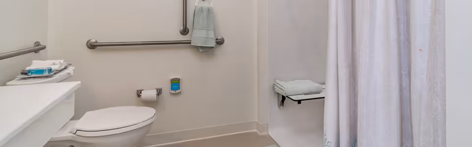 A clean and accessible bathroom featuring a white toilet with a closed lid, grab bars on the walls for support, a toilet paper holder with a roll of toilet paper, a towel hanging on a grab bar, a shelf with folded towels, and a shower area with a white patterned shower curtain.