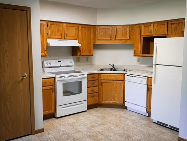 A kitchen with wooden cabinets, a white electric stove with oven, a white dishwasher, a white refrigerator, a double sink, and beige tiled flooring. There is a wooden door on the left side of the image.