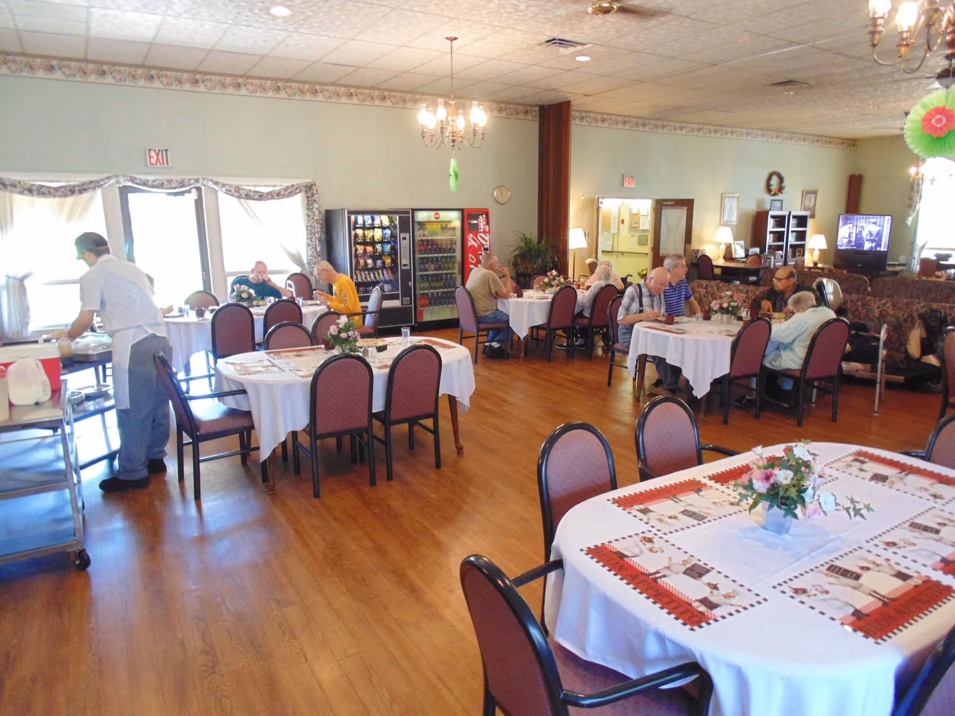 A dining room in a senior living facility with several round tables covered with white tablecloths and floral centerpieces. Elderly residents are seated at some tables eating and conversing. A staff member is serving food from a cart. There are vending machines against the far wall and a television in the background. The room has wooden flooring and soft lighting from chandeliers and lamps.