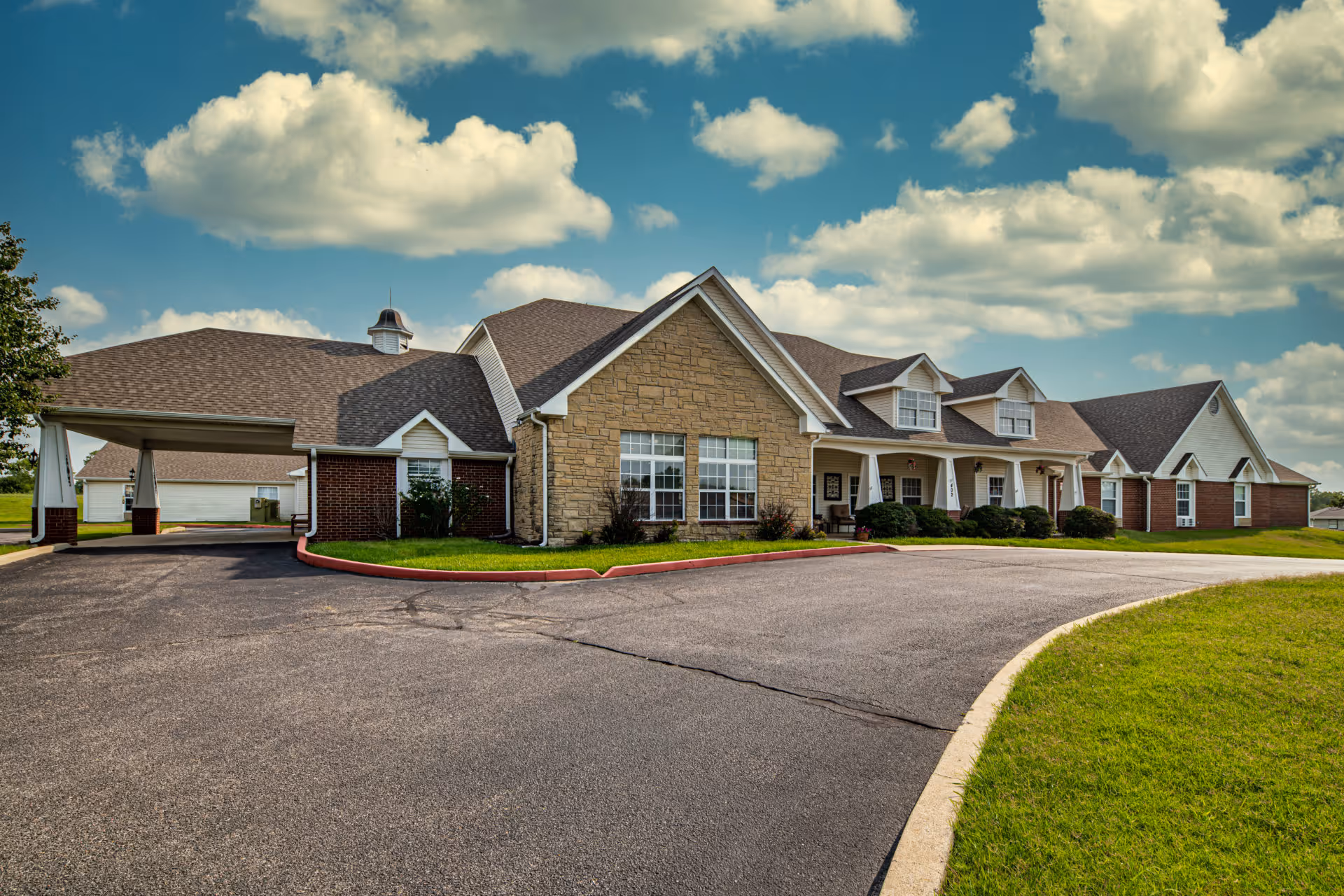 Exterior view of a single-story senior living facility building with a combination of brick and stone facade, multiple windows, a covered entrance driveway, and a well-maintained lawn under a partly cloudy sky.