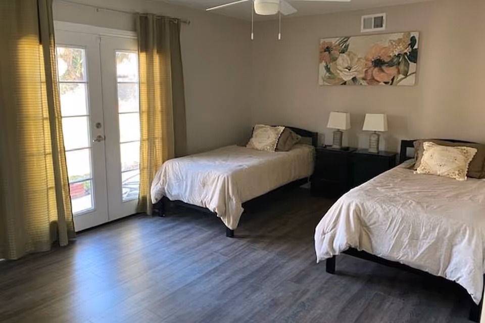A bedroom with two single beds, each with beige bedding and decorative pillows. Between the beds is a dark nightstand with two matching lamps. A floral painting hangs on the wall above the nightstand. The room has wooden flooring and a set of glass double doors with green curtains allowing natural light to enter.