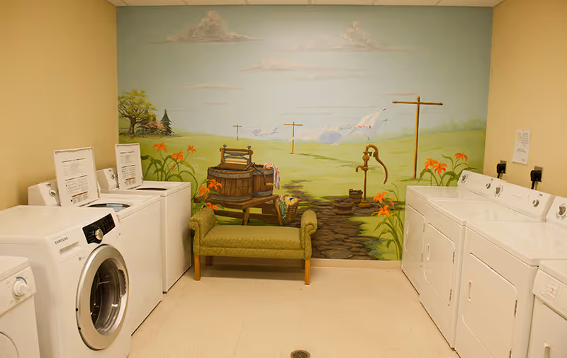 A laundry room with washers and dryers on either side and a green bench in front of a pastoral mural on the back wall.