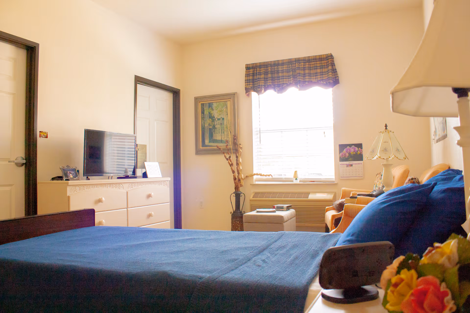 A cozy bedroom in an assisted living facility with a bed covered in a blue blanket in the foreground. There is a white dresser with a TV on top against the wall, a window with a plaid valance letting in natural light, and a small sitting area with two orange chairs and a lamp. A framed painting and a calendar are on the walls, and a vase with decorative branches is placed near the window.