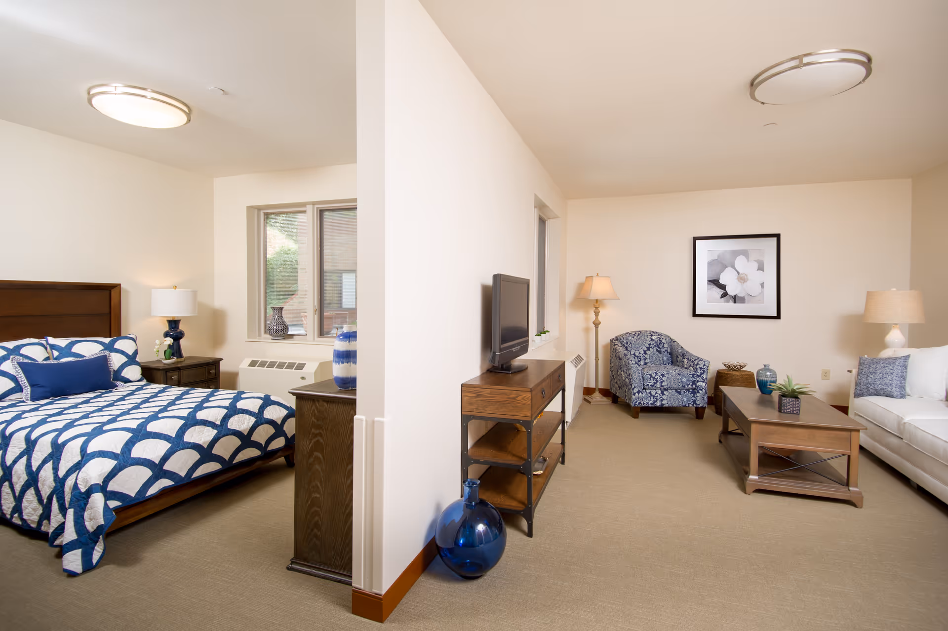 A bright and spacious senior living apartment featuring a bedroom with a wooden bed frame, blue and white patterned bedding, a nightstand with a lamp, and a window. Adjacent to the bedroom is a living room area with a wooden coffee table, a white sofa, a blue patterned armchair, a floor lamp, a TV on a wooden stand, and decorative items including vases and a framed flower artwork on the wall.