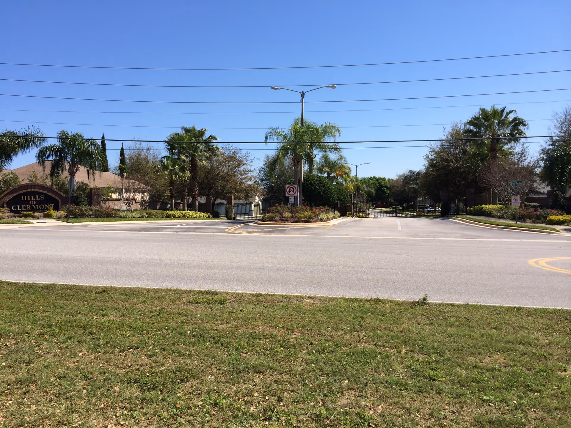 Gated community entrance with a road, palm trees, and a 'Hills of Clermont' sign under a clear blue sky.