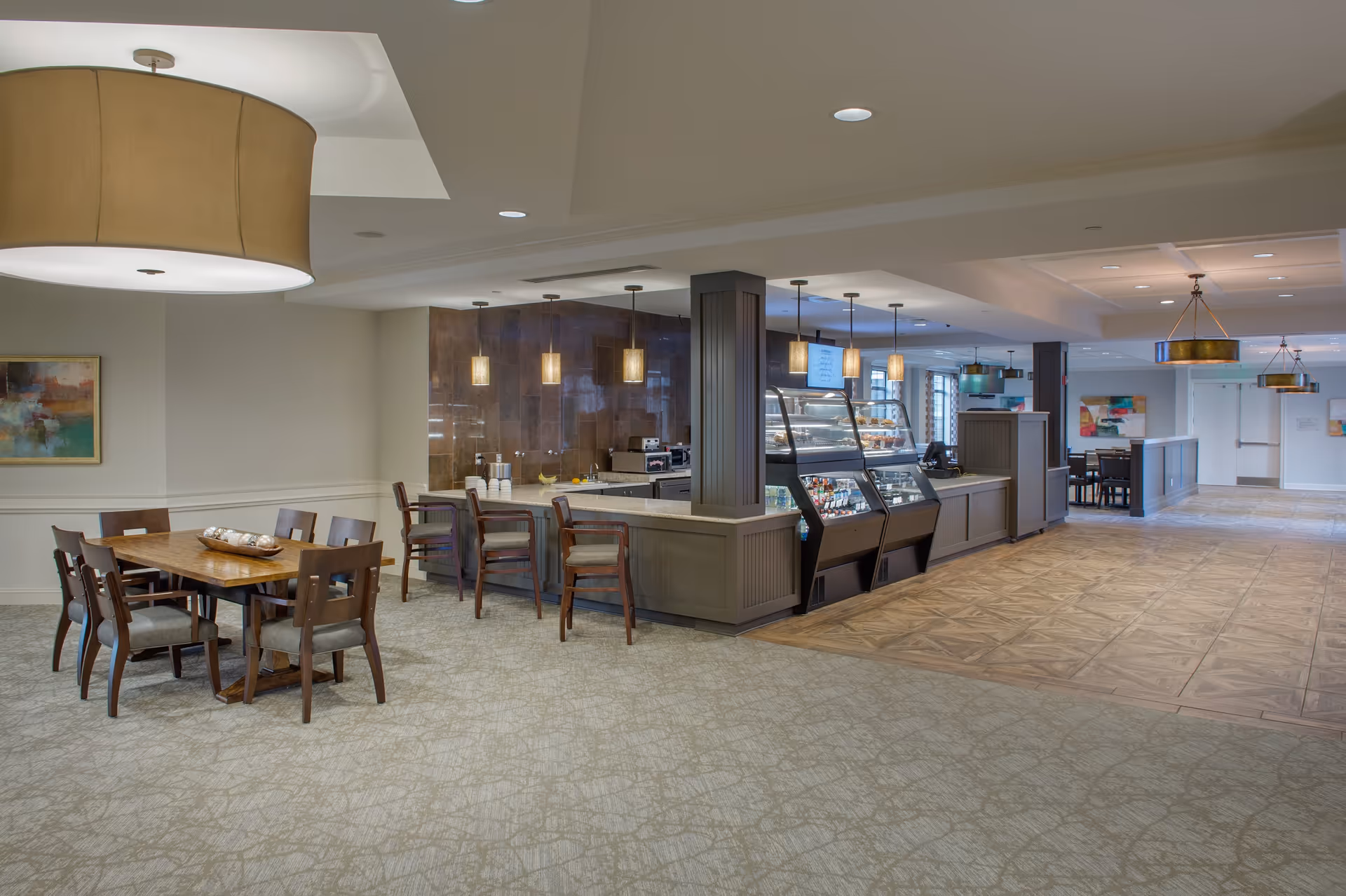 Interior view of a senior living facility dining and cafe area with a wooden dining table and chairs on the left, a counter with display cases for food and beverages in the center, and additional seating and lighting fixtures in the background.