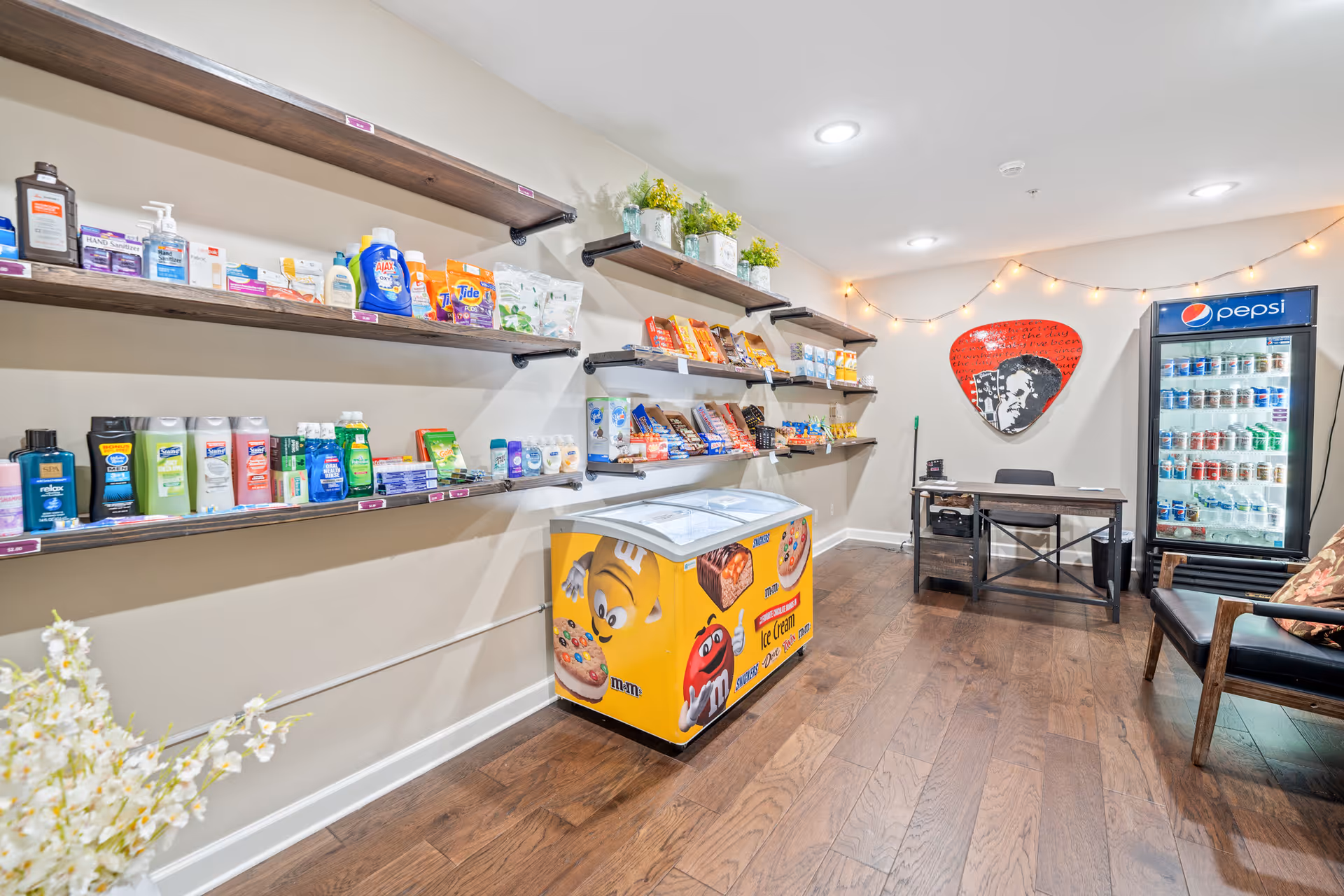 A small convenience store area inside a senior living facility with shelves stocked with toiletries, snacks, and household items. There is a yellow ice cream freezer with M&M's branding, a Pepsi refrigerator filled with beverages, a desk with a chair, and a wooden chair with a floral cushion. The walls are decorated with string lights and a red guitar pick-shaped wall art.