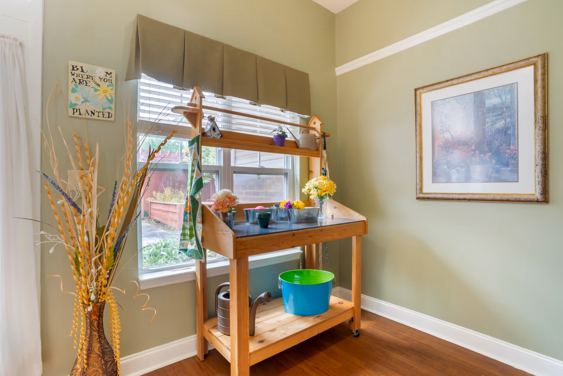 A bright indoor gardening station with a wooden potting bench in front of a window with white blinds and a green valance. The bench holds small flower pots, a watering can, and a blue bucket underneath. To the left is a tall decorative vase with dried plants, and on the wall above the vase is a sign that reads 'Bloom where you are planted.' On the right wall, there is a framed picture of a garden scene.