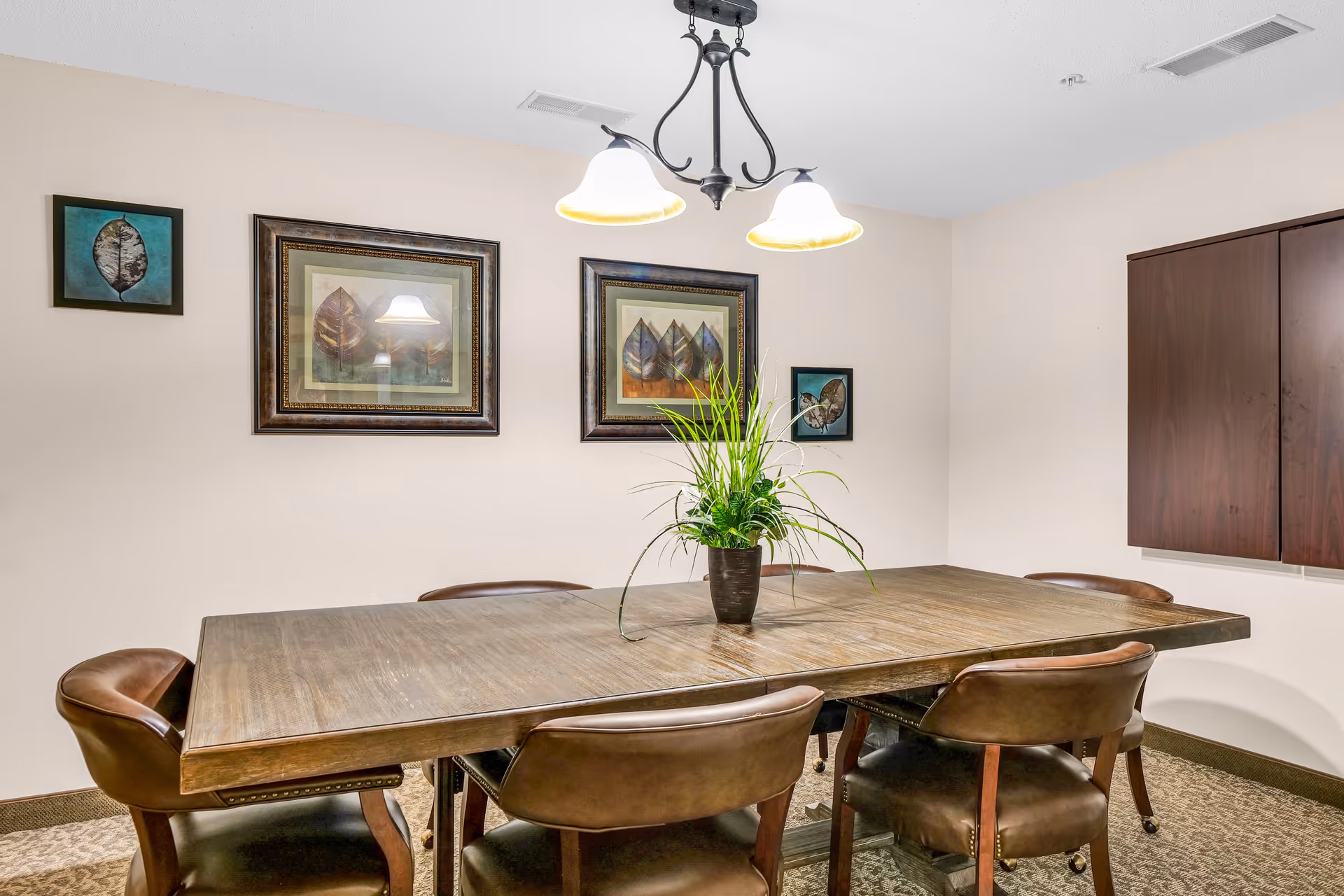 A dining room with a rectangular wooden table surrounded by six brown leather chairs. A green potted plant is centered on the table. The walls are decorated with framed artwork featuring leaves. A ceiling light fixture with two lamps hangs above the table.