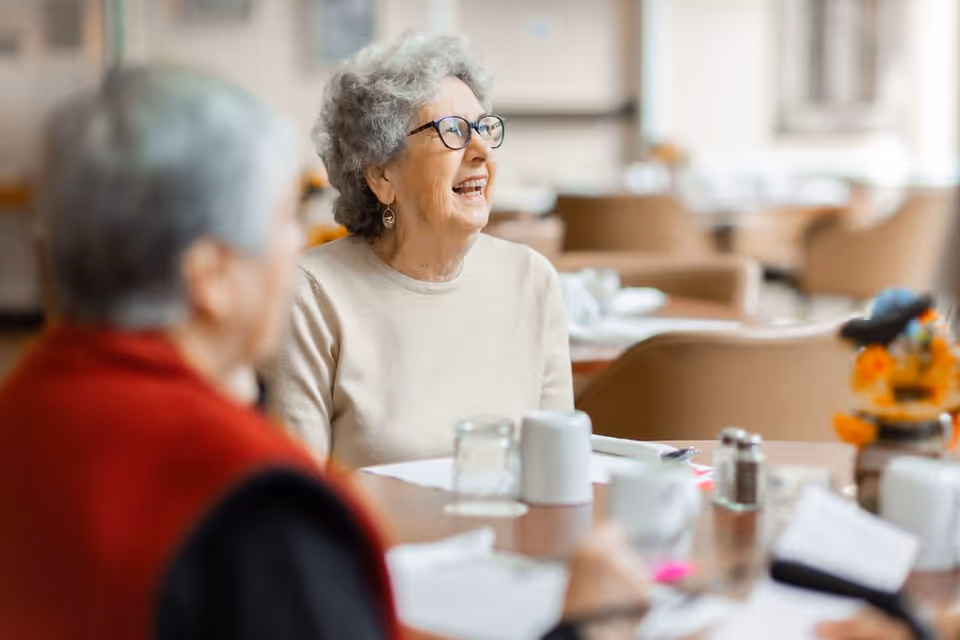 Two elderly women sitting at a dining table in a well-lit room, one woman with gray curly hair and glasses smiling and looking to the side, with cups, glasses, and condiments on the table.