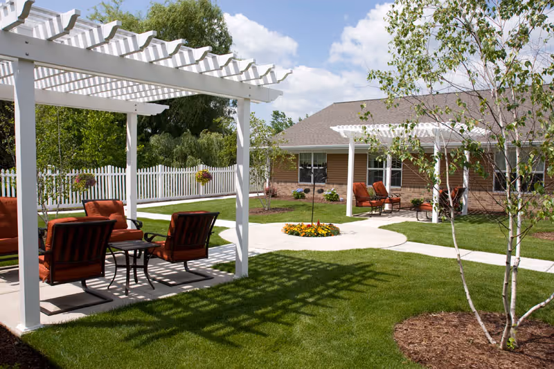 Sunny outdoor courtyard with white pergolas, red-cushioned patio chairs, paved walkways, manicured lawn and a one-story building in the background.