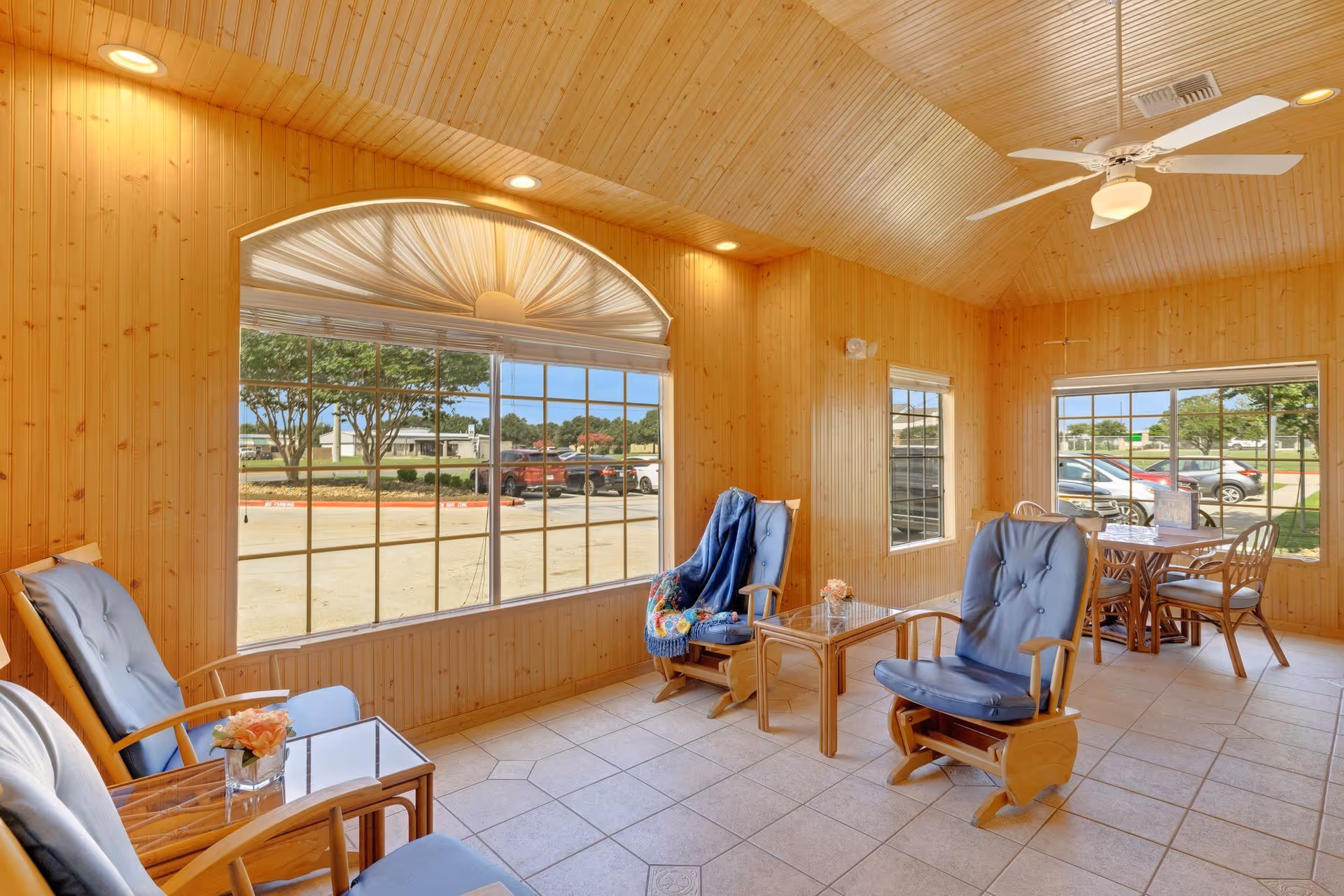 Sunlit wood-paneled common room with cushioned rocking chairs, small tables and a dining area by large windows overlooking the parking lot.