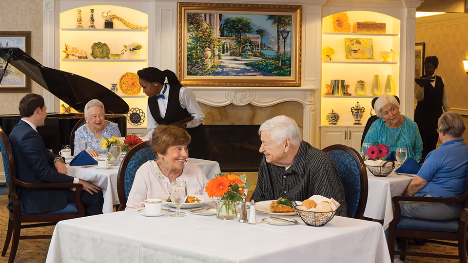 Several elderly residents and a younger man are seated at tables in a warmly lit dining room with white tablecloths, flowers, and place settings. Two waitstaff in black vests and white shirts are serving the tables. The room features a grand piano, built-in shelves with decorative items, and a large framed painting above a fireplace.