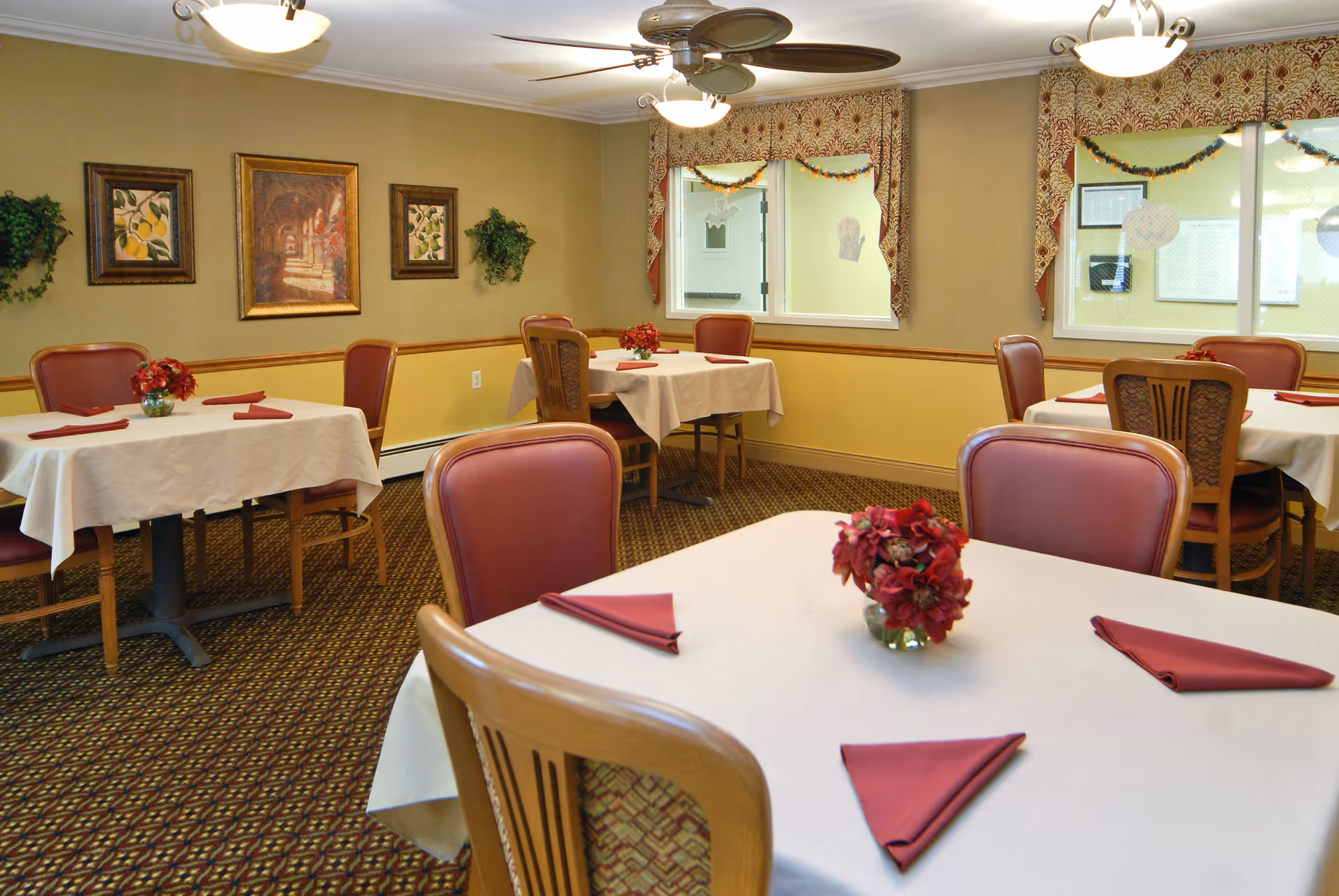 Well-lit dining room with multiple tables set with white tablecloths, red napkins and floral centerpieces, surrounded by upholstered chairs and wall artwork.
