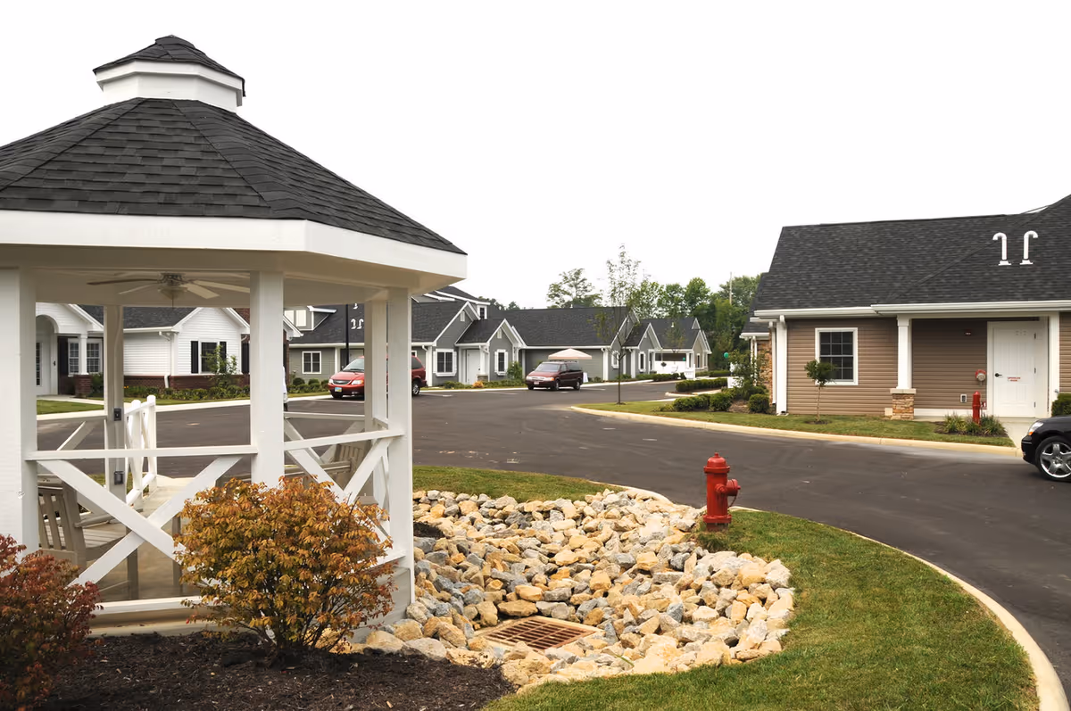 Outdoor view of a senior living community featuring a white gazebo with a shingled roof and ceiling fan, surrounded by landscaping with bushes and rocks. In the background, there are several single-story residential buildings with pitched roofs, a paved road, parked cars, and a red fire hydrant near the curb.
