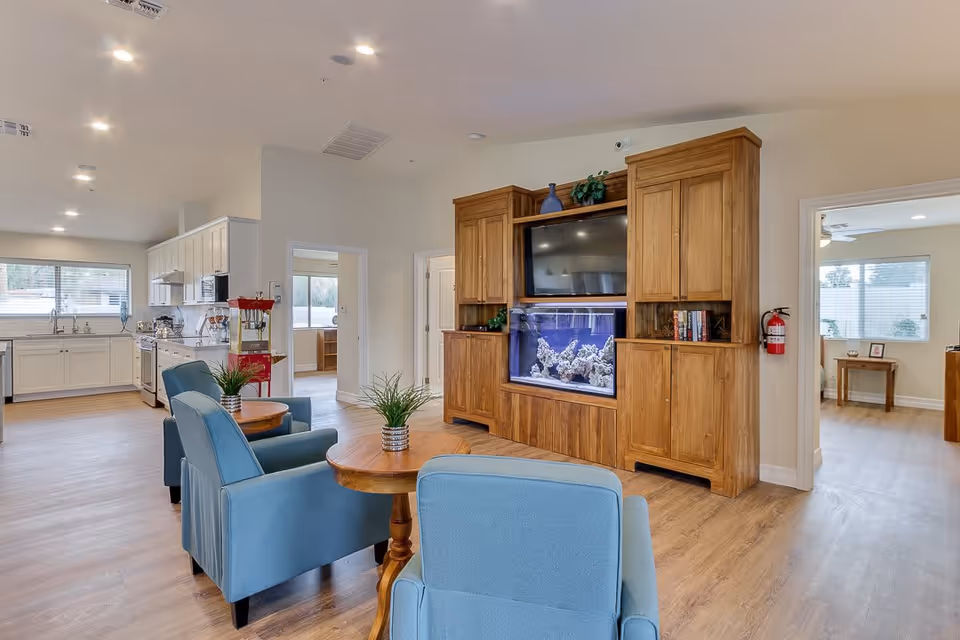 A bright and spacious common area in a senior living facility featuring light wood flooring, blue upholstered chairs around small wooden tables with potted plants, a large wooden entertainment center with a built-in aquarium and a flat-screen TV, and a kitchen area with white cabinets and stainless steel appliances in the background.