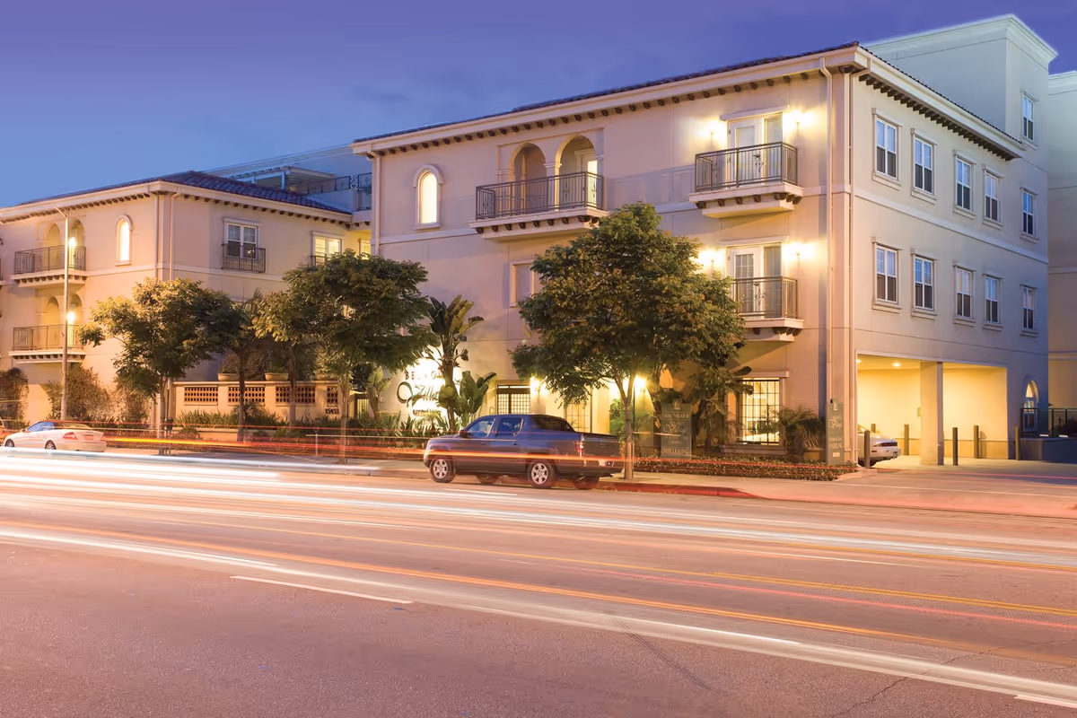 Exterior view of Belmont Village Senior Living Encino building at dusk with illuminated windows, trees, parked cars, and light trails from passing vehicles on the street in front.