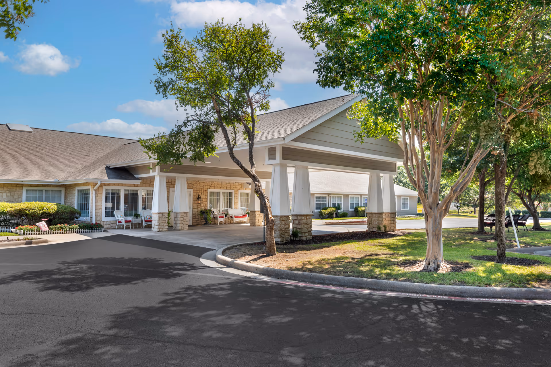 Exterior view of Brookdale White Rock senior living facility entrance with a covered driveway supported by white pillars with stone bases, surrounded by trees and greenery under a partly cloudy sky.