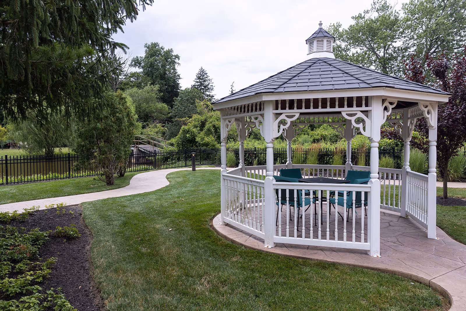A white wooden gazebo with a shingled roof situated on a paved area surrounded by green grass and landscaped garden beds. There are teal chairs and a table inside the gazebo. A curved concrete pathway leads past the gazebo, with trees and shrubs in the background under a cloudy sky.