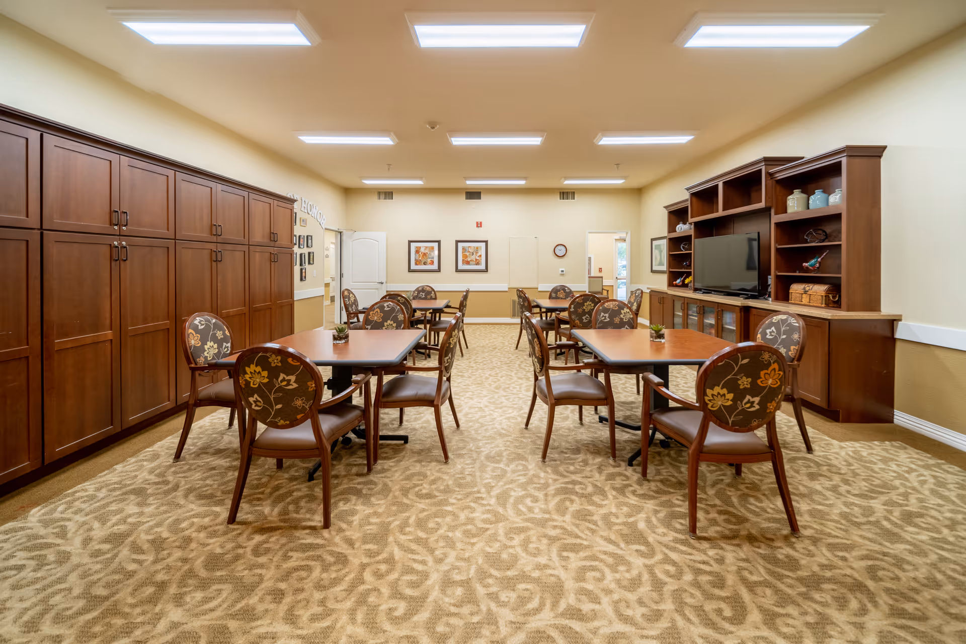 A well-lit room with multiple square wooden tables and floral-patterned chairs arranged neatly on a beige patterned carpet. The room features wooden cabinets along the left wall and a large wooden entertainment center with a flat-screen TV and decorative items on the right wall. The walls are painted light yellow with framed artwork and a clock visible in the background.