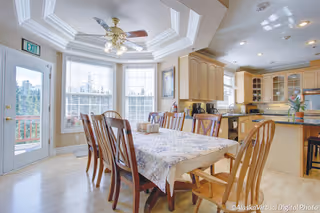 Bright dining area with a wooden table covered by a floral tablecloth surrounded by eight wooden chairs. The room features large windows with white blinds letting in natural light, a ceiling fan with lights, and light-colored cabinetry in the adjacent kitchen area. A door with an exit sign leads outside to a deck.