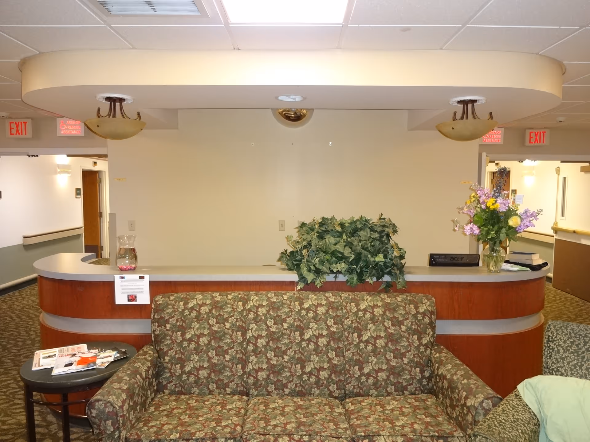 Reception area in a senior living facility with a floral patterned couch in front of a curved wooden desk. The desk has a vase with flowers, a green leafy plant, a pitcher of water, and some papers. There are two ceiling lights above the desk and exit signs visible in the background hallway.