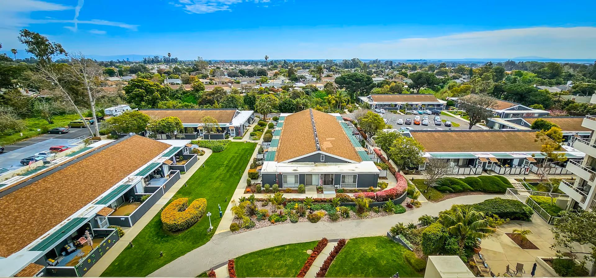 Aerial view of Ventura Townehouse senior living facility showing multiple single-story buildings with brown roofs, well-maintained green lawns, landscaped gardens, pathways, and a parking area in the background under a clear blue sky.