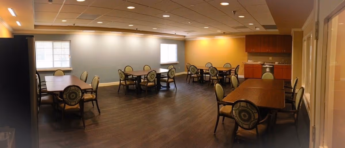 Large common dining room with several tables and upholstered chairs, wood-look flooring, windows, and a kitchenette.