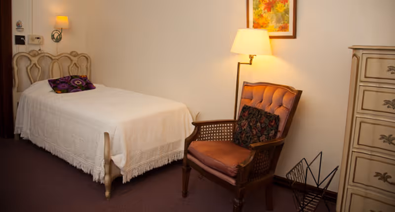 A small bedroom with a white-covered twin bed, upholstered armchair, floor lamp, and a dresser.