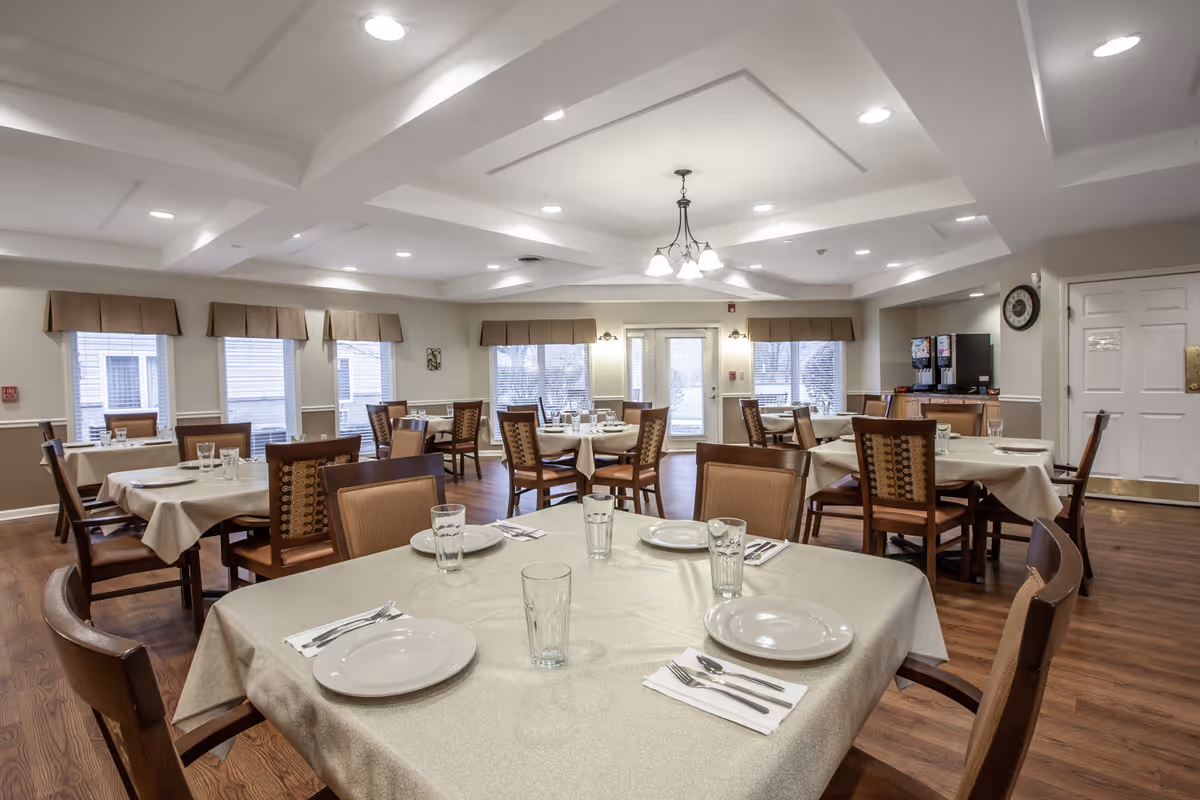 A dining room in a senior living facility with multiple tables covered in beige tablecloths, each set with plates, glasses, and silverware. The room has wooden chairs, large windows with brown valances, a chandelier, and a beverage station in the corner.