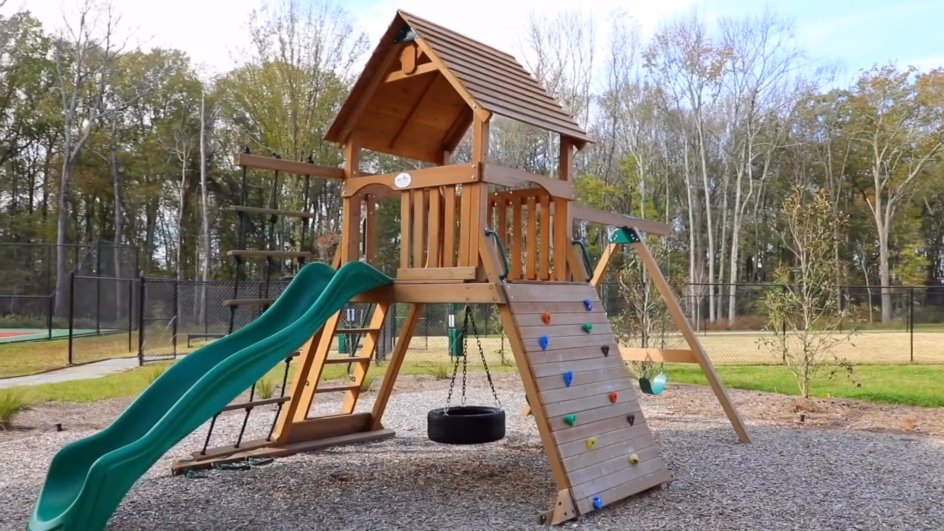 Outdoor playground structure with a green slide, climbing wall with colorful grips, tire swing, and a small wooden playhouse with a roof, set on a gravel surface with trees and a fenced area in the background.