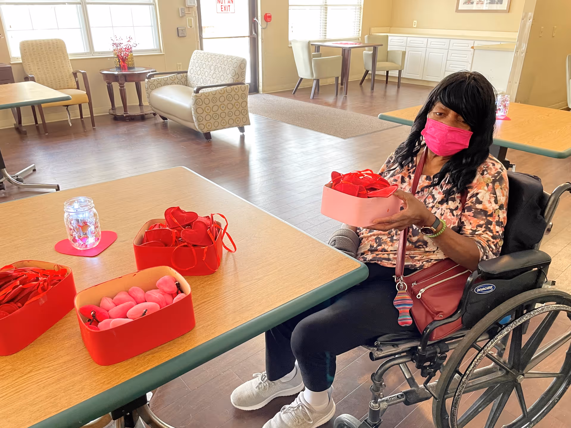 A person in a wheelchair wearing a pink face mask holds a pink box filled with red and pink heart-shaped items while seated at a table in a bright common/dining room.
