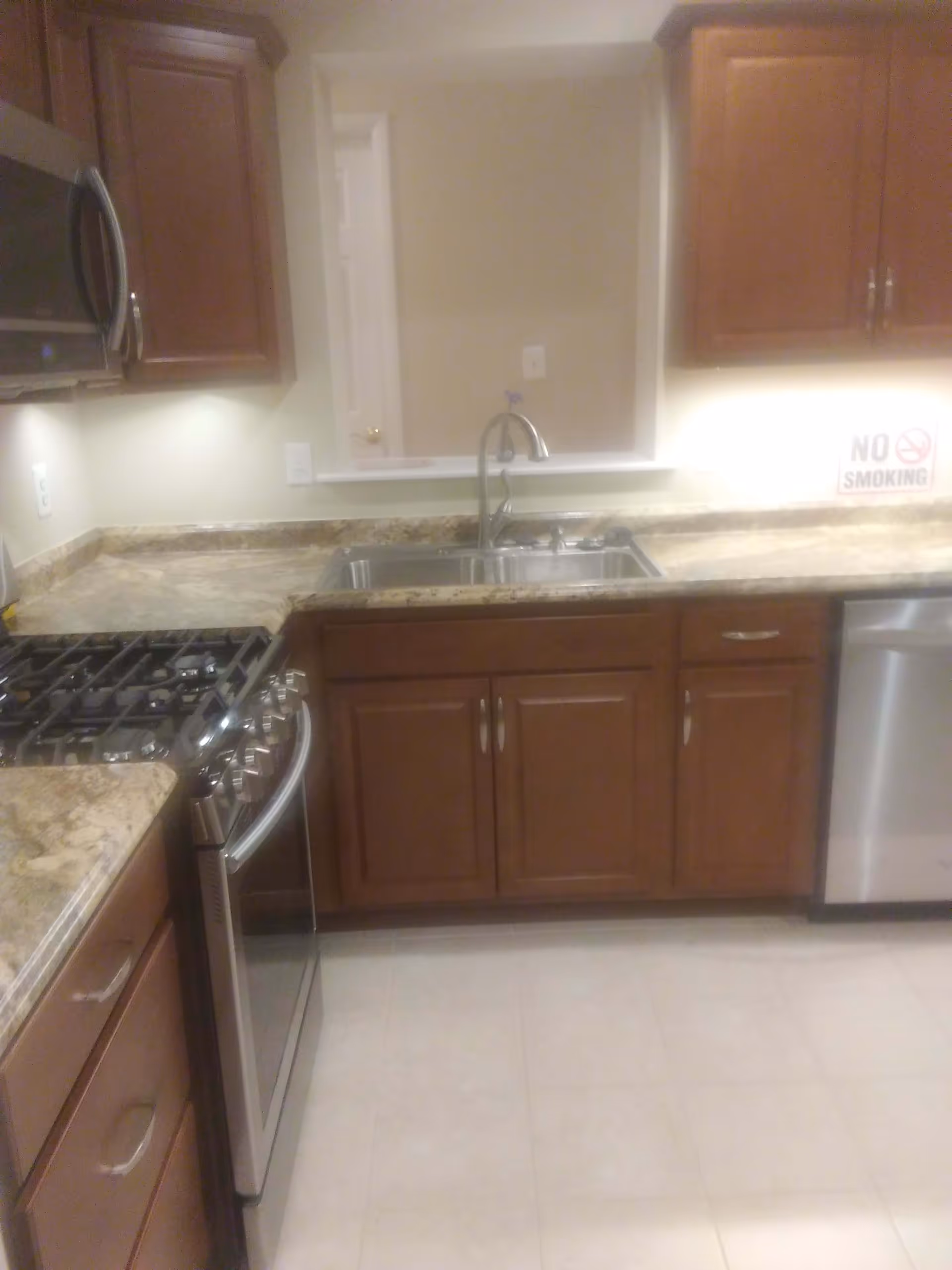 A kitchen with wooden cabinets, a granite countertop, a stainless steel gas stove, a microwave, a double sink with a faucet, and a dishwasher. There is a 'No Smoking' sign on the countertop near the sink.