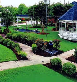 A well-maintained outdoor garden area with green grass, trimmed bushes, a paved walkway, benches, a white gazebo, and a bird feeder on a pole surrounded by trees and shrubs.