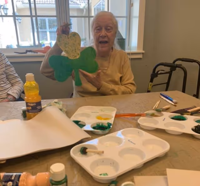An elderly woman sitting at a table in a room with large windows, holding up a painted shamrock craft. The table is covered with art supplies including paint palettes, brushes, and bottles of paint. A walker is visible in the background.