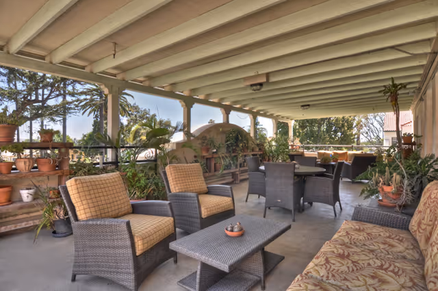 Covered outdoor patio area with wicker chairs and a table, surrounded by potted plants and greenery, with a ceiling fan and beams overhead.