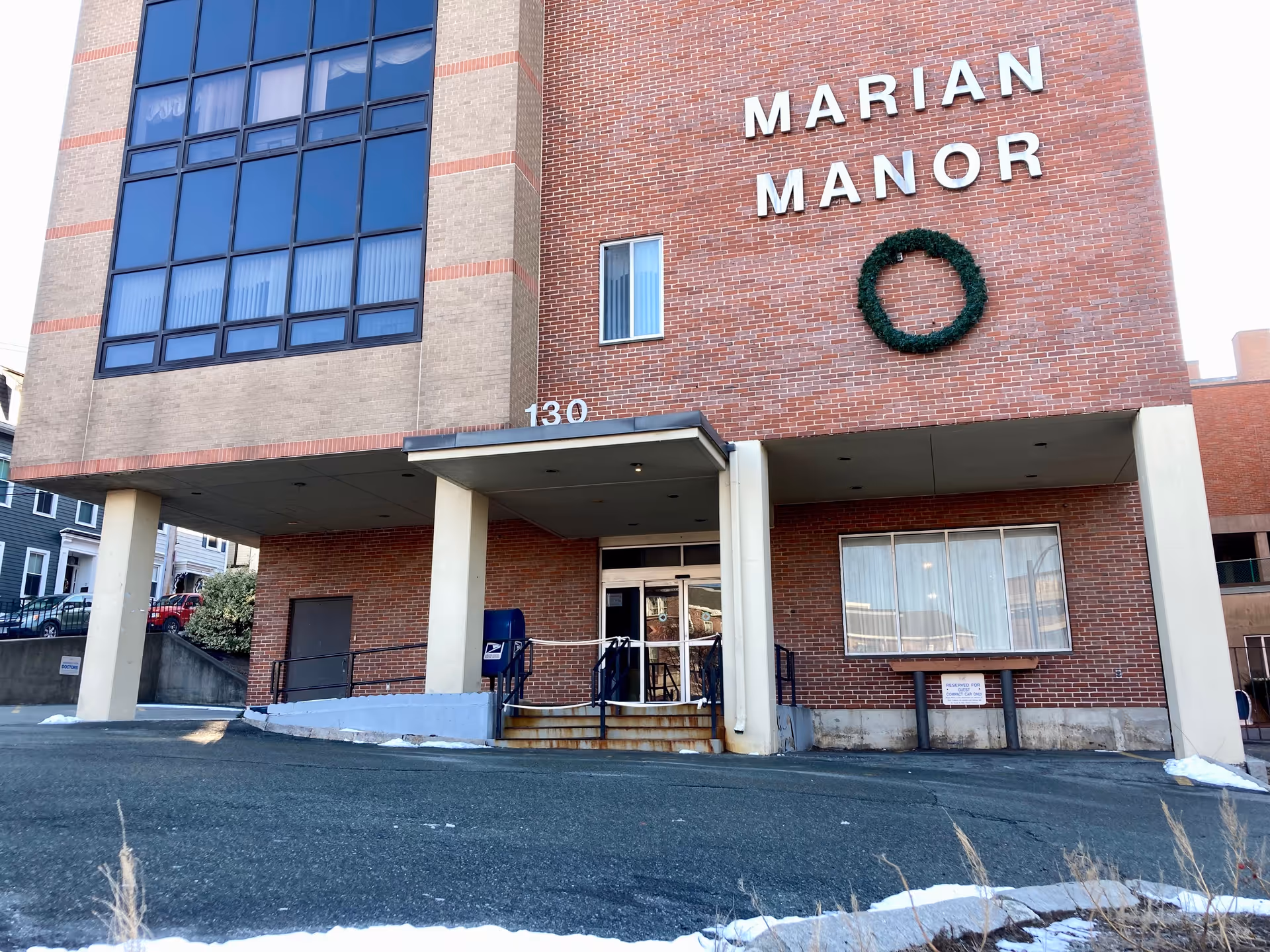 Exterior view of Marian Manor Nursing Home building with a brick facade, large windows, and an entrance with stairs and a ramp. A green wreath is mounted on the wall near the building name.