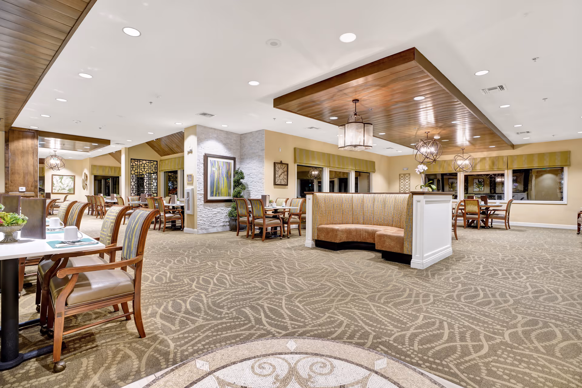 Spacious dining area with multiple wooden tables and chairs arranged neatly. The room features a patterned carpet, warm lighting with decorative ceiling fixtures, and a curved booth seating area in the center. Walls are painted in neutral tones with framed artwork and large windows with valances.