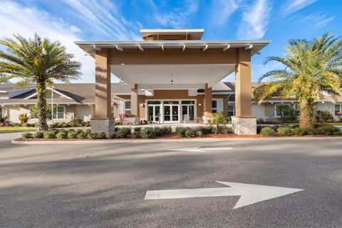 Front exterior view of a senior living facility with a covered entrance supported by columns, landscaped with palm trees and shrubs, under a blue sky with some clouds.