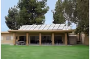 Single-story building with a covered patio area featuring tables and chairs, surrounded by green grass and tall trees in the background under a clear sky.