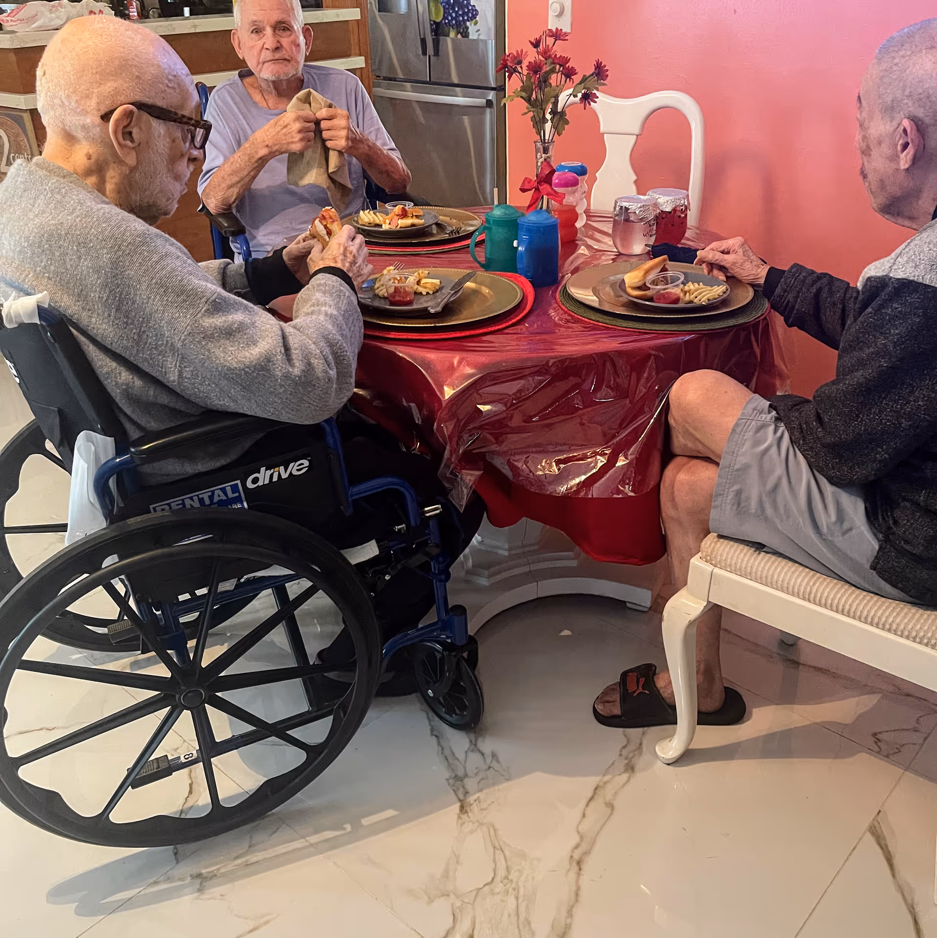 Three elderly men sitting around a round dining table covered with a red tablecloth, eating a meal. One man is in a wheelchair, and the other two are seated on chairs. The table has plates with food, cups, and a small vase with flowers. The background shows a kitchen area with a refrigerator and cabinets.