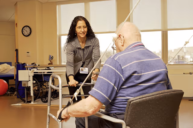A woman with curly hair and a gray jacket assists an elderly man in a blue striped shirt using a walker in a rehabilitation or therapy room with exercise equipment and large windows in the background.
