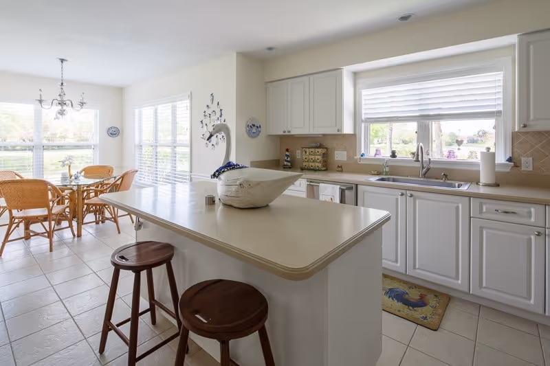 Bright open kitchen with white cabinets, a center island with two wooden stools, and a dining area by large windows.