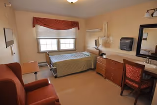 A furnished single-occupancy bedroom with a bed, recliner, dresser, sink, and window with valance.