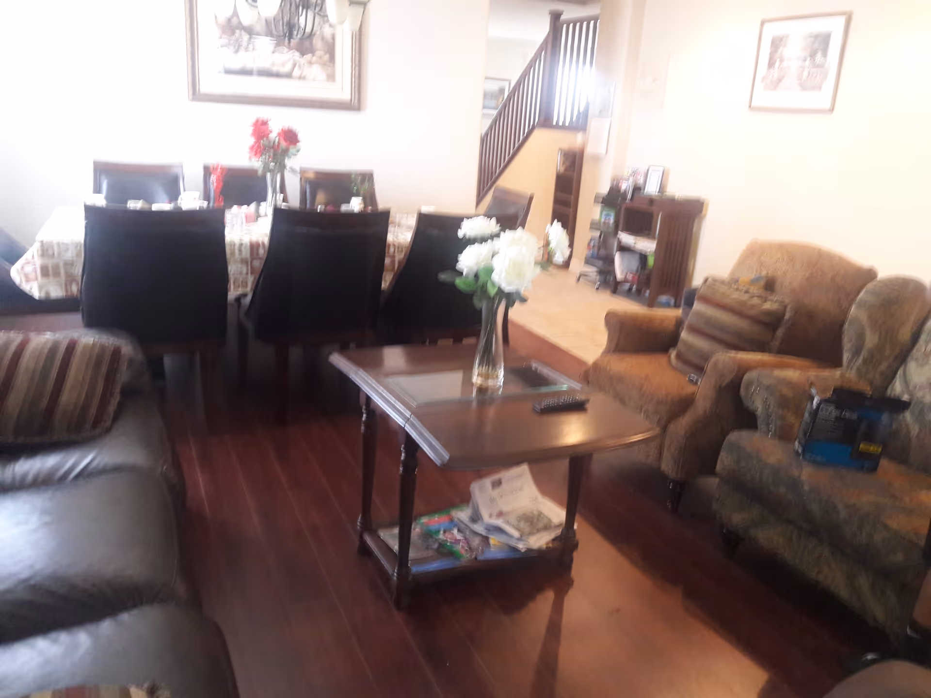 A cozy living room and dining area with wooden flooring. The living room has a brown armchair with striped cushions, a patterned armchair, and a leather sofa. A wooden coffee table with a glass top holds a vase of white flowers and some magazines underneath. The dining area features a table covered with a patterned tablecloth and surrounded by black chairs. A staircase and a small wooden cabinet are visible in the background.
