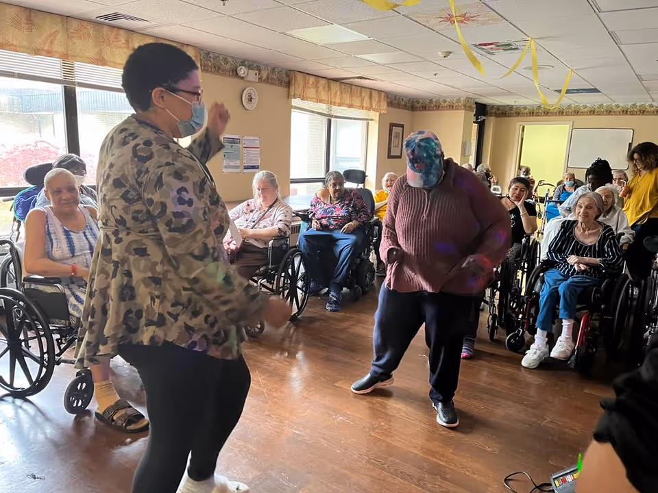 A group of elderly people, many in wheelchairs, are gathered in a room with large windows. Two individuals are standing and appear to be dancing or leading an activity, while others watch and smile. The room has wooden flooring and light-colored walls with floral border wallpaper near the ceiling.