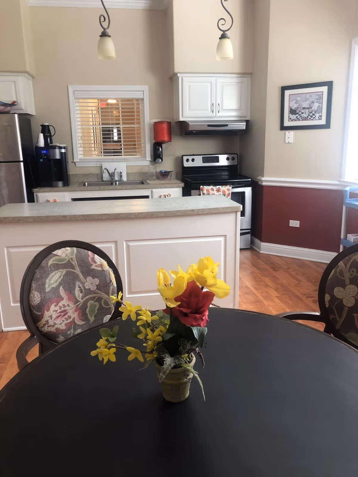 Interior view of a kitchen area in a senior living facility with a round black table in the foreground holding a small vase of yellow and red artificial flowers. Behind the table is a kitchen counter with a sink, stainless steel refrigerator, coffee maker, stove, and white cabinets. The walls are beige with a maroon lower section and there is a framed picture on the wall to the right.