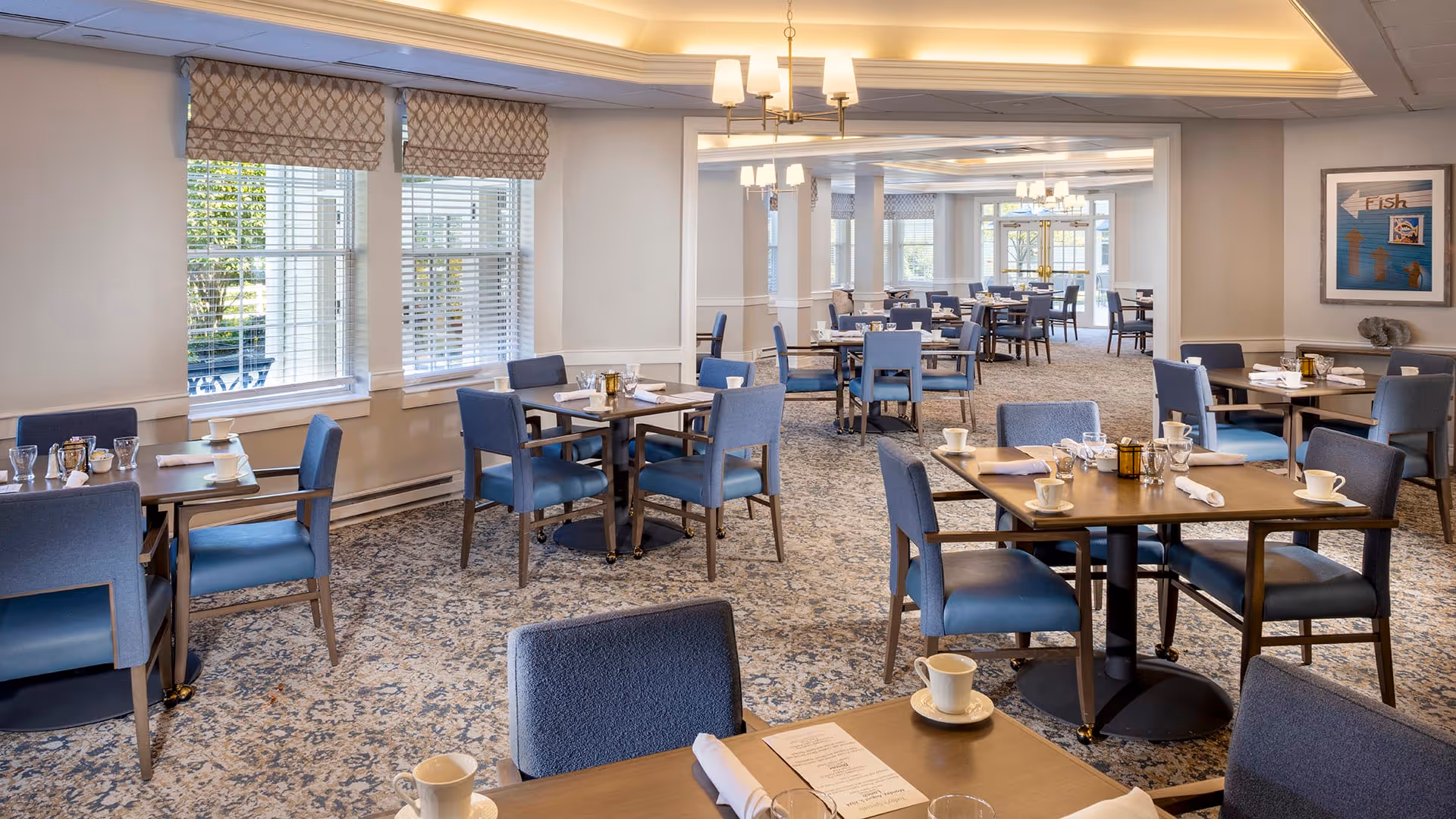A spacious dining room with multiple wooden tables and blue upholstered chairs. The tables are set with white cups, glasses, napkins, and menus. Large windows with patterned valances allow natural light to fill the room. The ceiling features recessed lighting and chandeliers, and the carpet has a blue and beige pattern. A framed artwork is visible on the wall to the right.