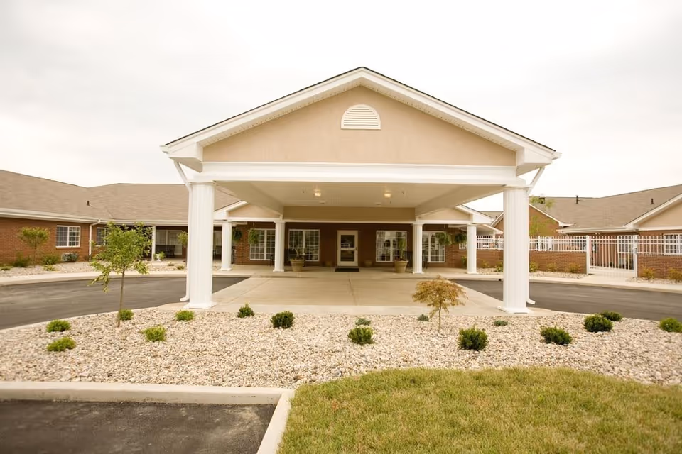 Front entrance of a single-story senior living building with a covered porte-cochere, white columns, and landscaped gravel beds.